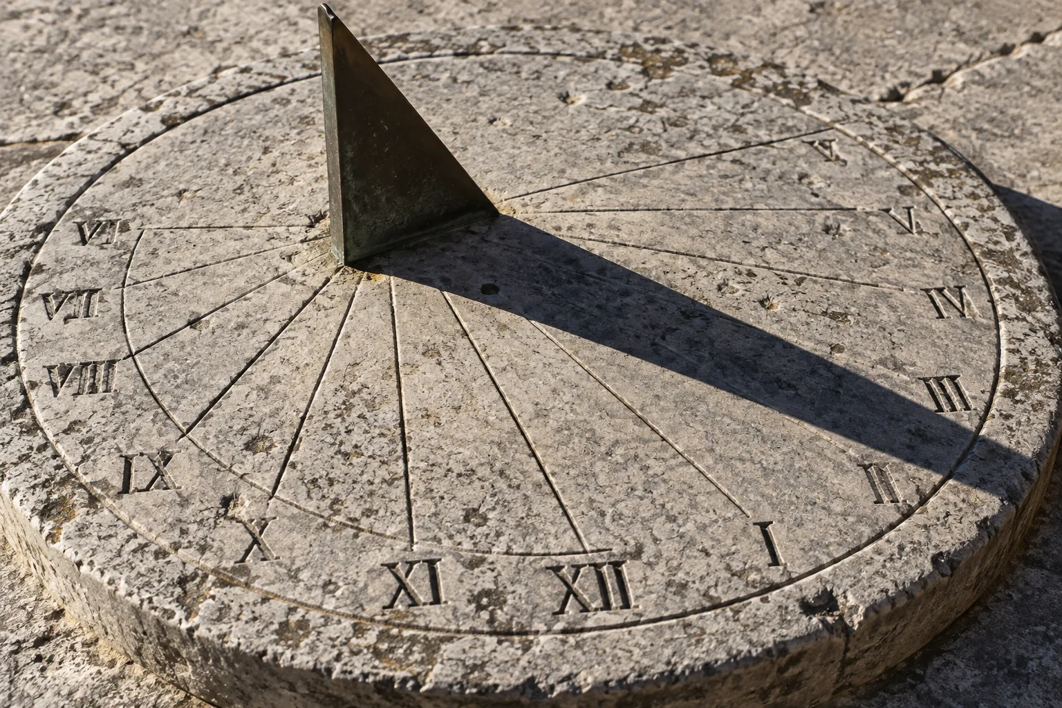 A weathered stone sundial with Roman numerals, its brass gnomon casting a hard diagonal shadow